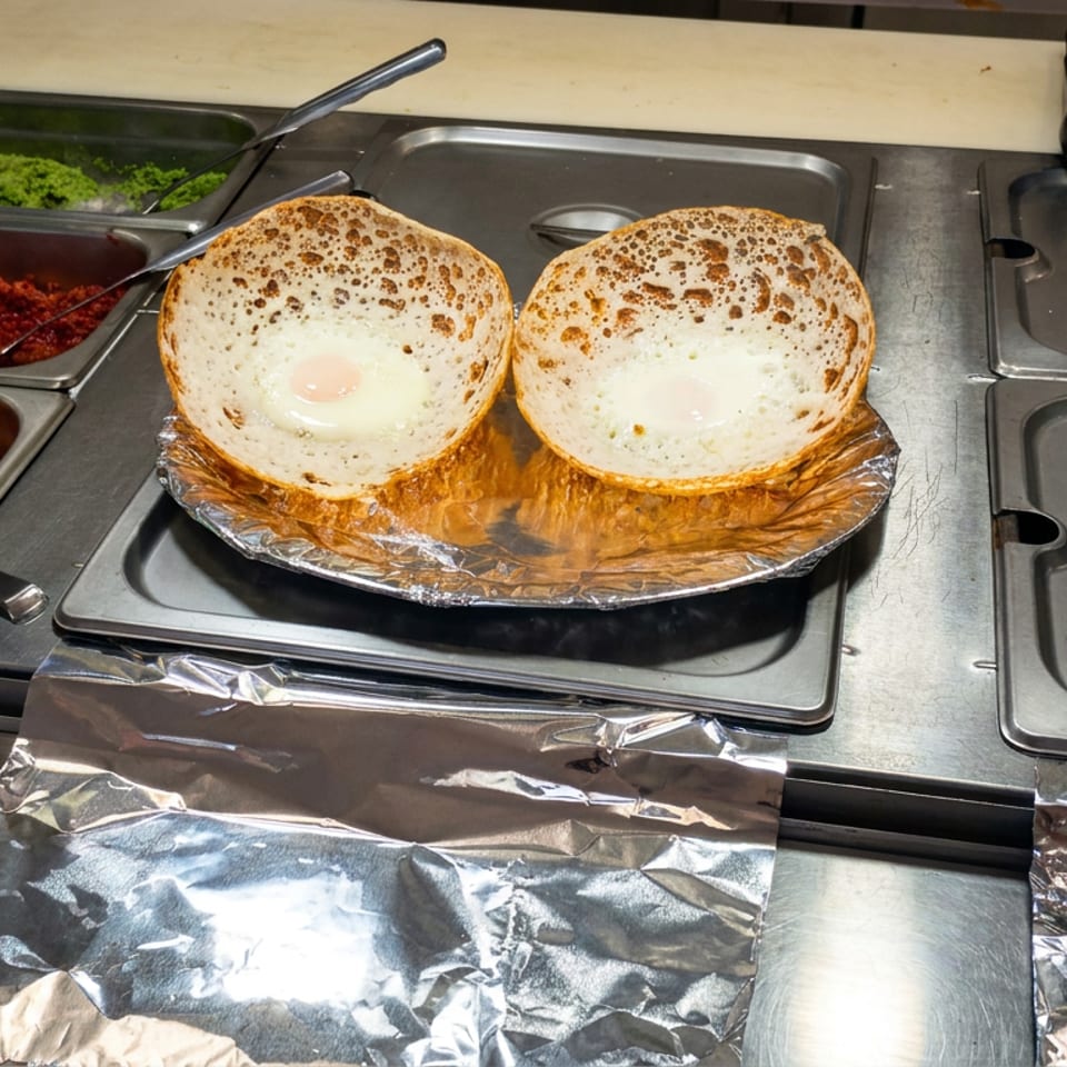 Two round toasted bread bowls holding eggs sit on foil at a restaurant buffet beside trays of colorful ingredients.