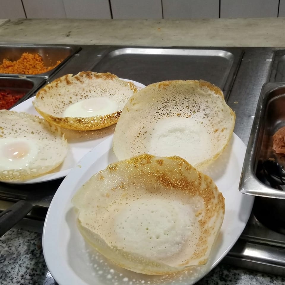 Four bowl-shaped pancakes, including an egg-topped pancake, sit on plates beside buffet trays of curries and noodles.