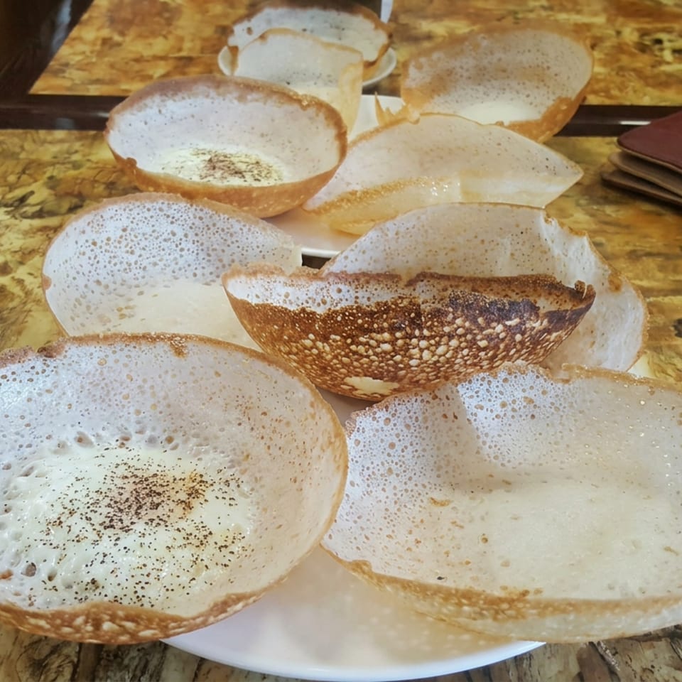 Plate of lacy, bowl-shaped South Indian appam pancakes on a marble restaurant table beside a leather wallet.